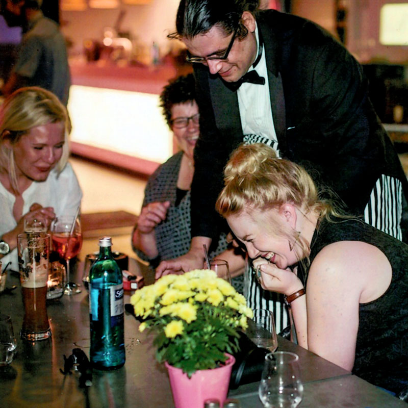 Gäste lachen herzlich während Spaßkellner Theo anlässlich eines Geburtstages an deren Tisch zaubert